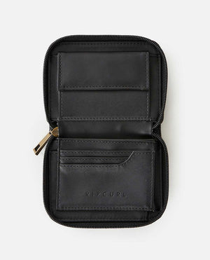 The Rip Curl Wanderer Small Zip Wallet, in black leather with "RIP CURL" embossed on the lower pocket, showcases minimalist design. It offers multiple card slots and a secure zippered edge, combining simplicity and functionality against a plain white background.