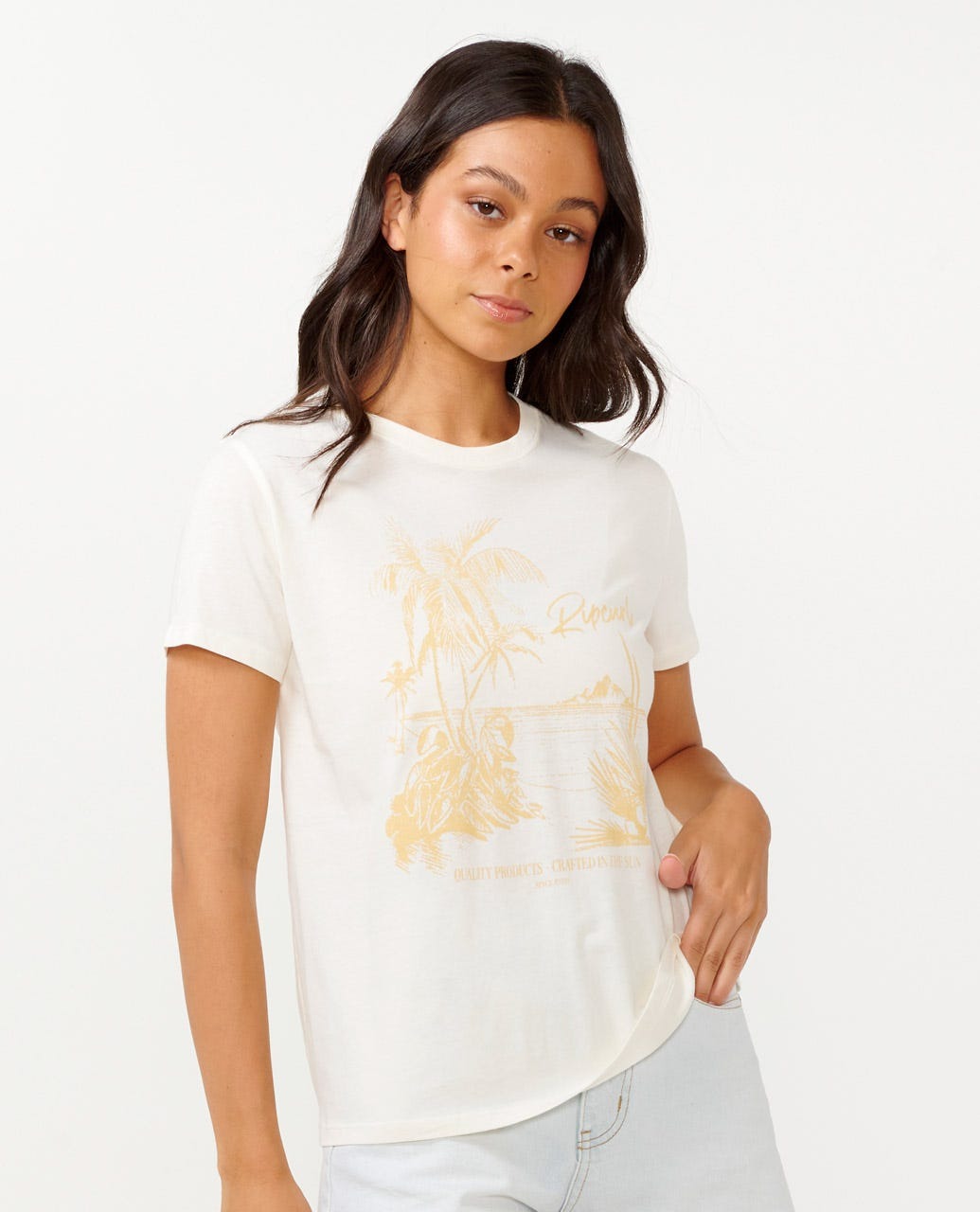 A person with long dark hair is wearing the Rip Curl Buena Vista Standard Tee, a white cotton jersey t-shirt featuring a palm tree design, paired with light blue jeans. They are posing against a plain backdrop.