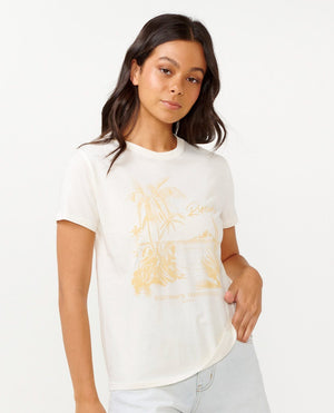 A person with long dark hair is wearing the Rip Curl Buena Vista Standard Tee, a white cotton jersey t-shirt featuring a palm tree design, paired with light blue jeans. They are posing against a plain backdrop.