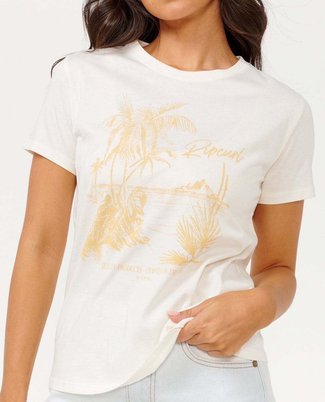 A person is wearing the Rip Curl Buena Vista Standard Tee, featuring a striking gold tropical print of palm trees and the phrase "Rip Curl Buena Vista" on a white T-shirt. The cotton jersey material ensures a comfortable, standard fit. Their hand is gently tucked into the shirt near the hem, set against a simple white backdrop.
