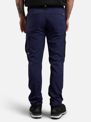 A person stands against a plain white background wearing navy King Gee Trademark Cargo Pants with black patches, black sneakers, and a black shirt. The ripstop fabric pants feature multiple pockets, and they have a tattoo on their left arm.