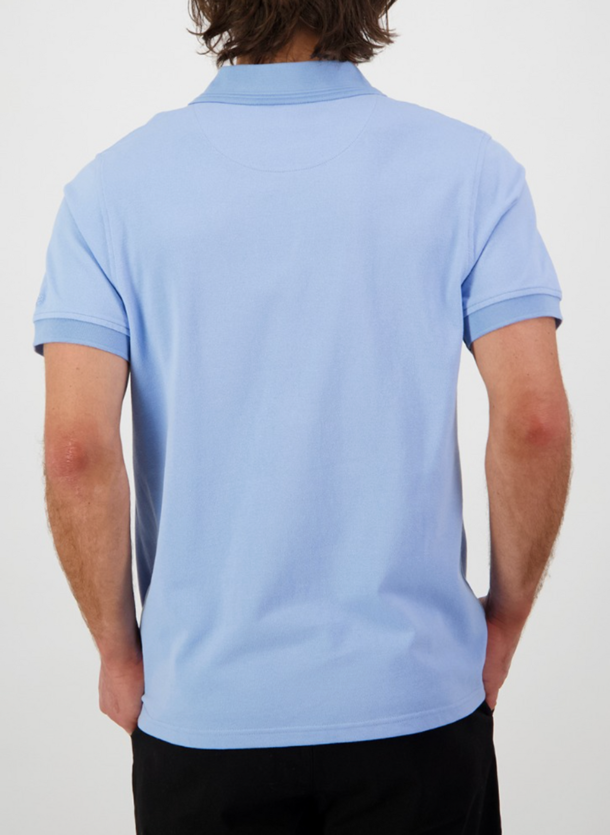 A person with medium-length brown hair wears a Swanndri Highgrove polo crafted from combed cotton pique and black pants, standing with their back to the camera against a plain white background.