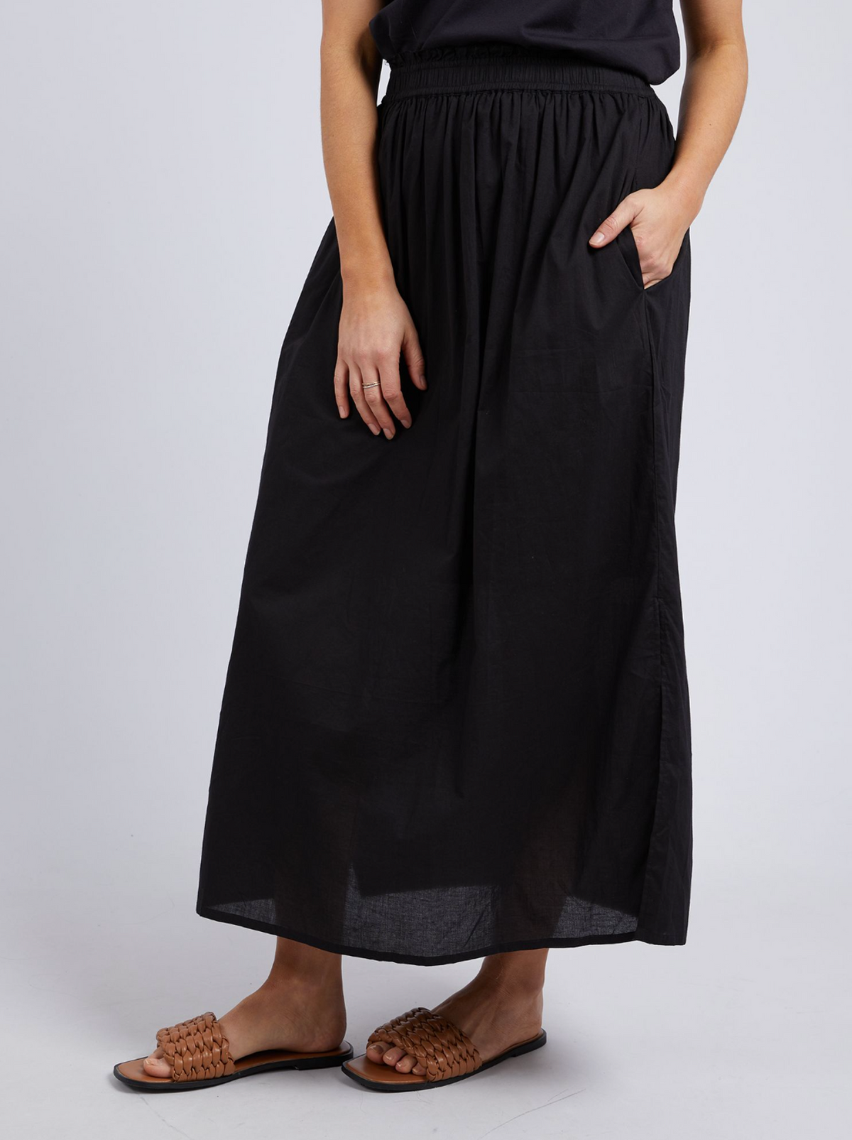 A person wearing a black, ankle-length Foxwood Charli Skirt with an elastic waist and pockets, paired with a black top and brown woven slide sandals, stands against a plain light background.
