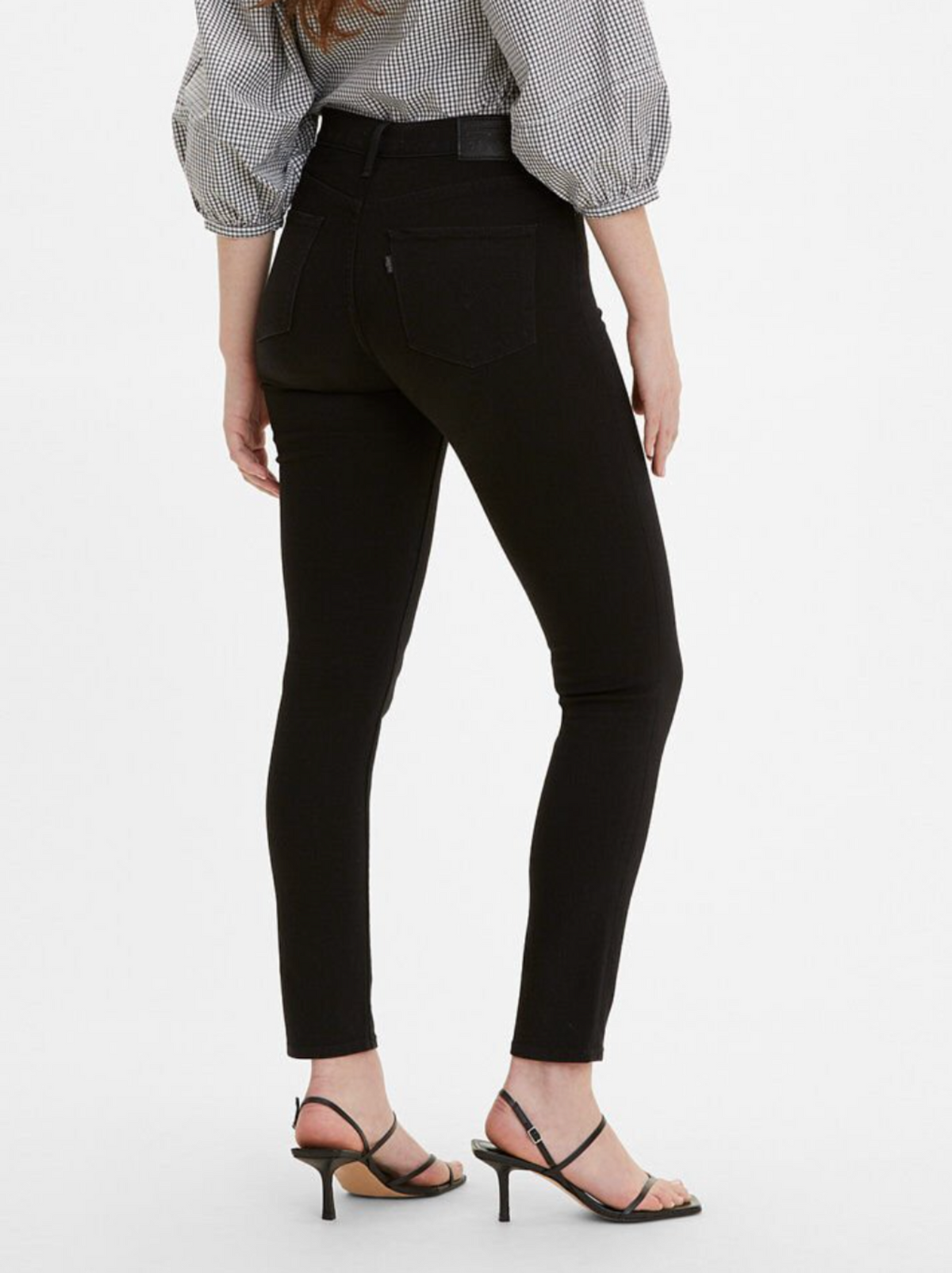 A person stands facing away from the camera, wearing Levis 311 Shaping Skinny Jean in black, a black-and-white checkered blouse with puffed sleeves, and black strappy high-heeled sandals. The person's hair is long and flowing down their back.