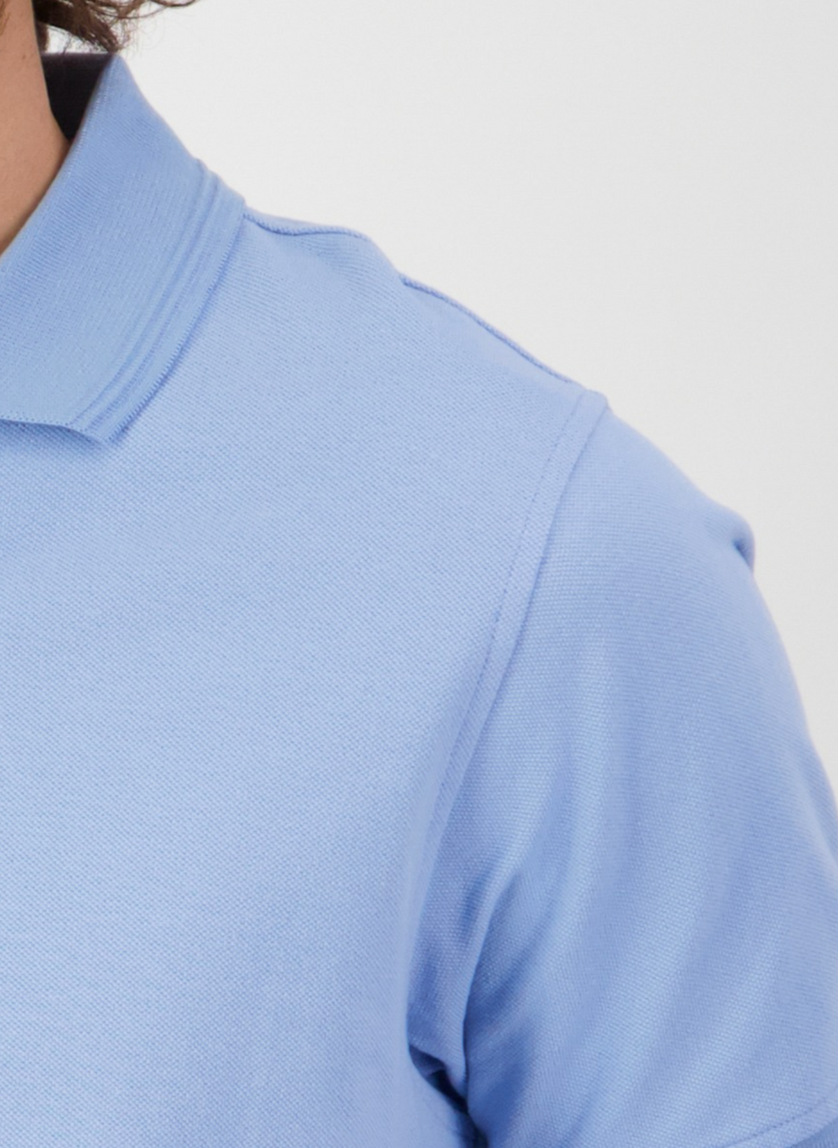 Close-up of an individual wearing a Swanndri Highgrove Polo in light blue, highlighting the upper portion made from combed cotton pique with detailed texture and stitching. The plain, bright background emphasizes the shirt's refined elegance.