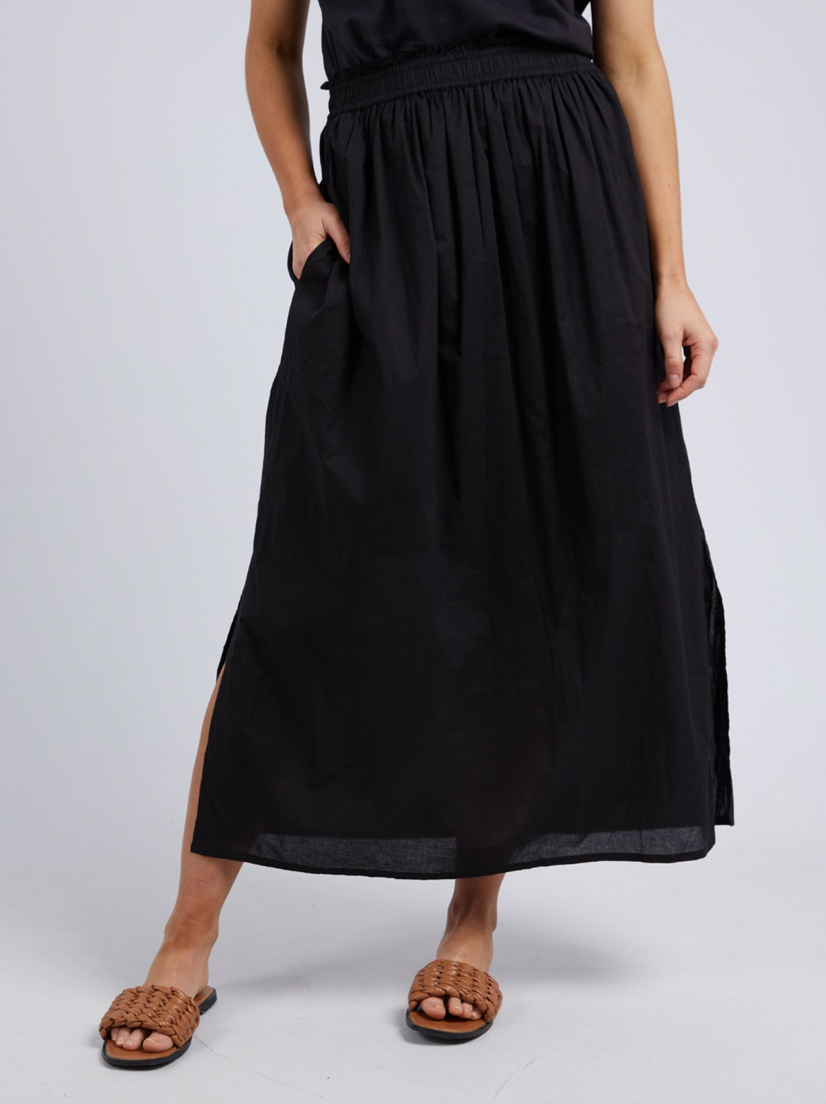 Wearing the Foxwood Charli Skirt—an ankle-length black 100% cotton skirt with side slits and an elastic waist—paired with a black top and brown woven sandals, a person stands with one hand in their pocket against a light background.