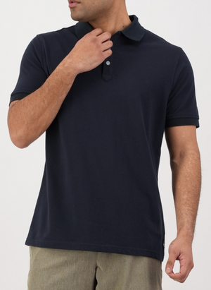 A man in a dark navy Swanndri Highgrove Polo and light beige pants stands against a plain background, touching his collar. His face is partially out of frame.