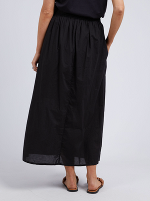 A person wearing the Foxwood Charli Skirt—a long black maxi with an elastic waist—paired with a black top and brown flat sandals stands with their back to the camera against a plain light gray background.