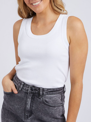 A person wearing a fashionable Foxwood Esme Tank and high-waisted, faded black jeans smiles with one hand in their pocket against a light-colored, plain background.