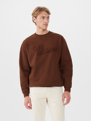 A man wearing a brown RM Williams R.M.W Script Crew Neck jumper and off-white pants stands against a plain white background, looking slightly to his left.