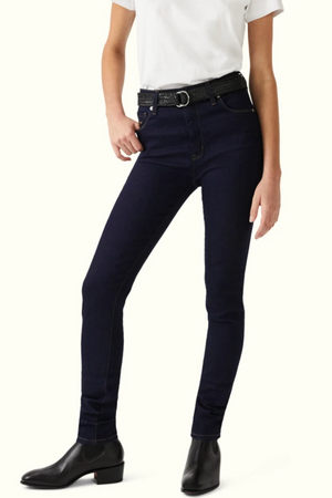 A person stands wearing a white shirt, dark blue RM Williams Albury skinny high waist jeans with a black belt, and black ankle boots. The high-rise skinny fit jeans feature front and back pockets, with one hand resting in a pocket and the other hanging by their side.