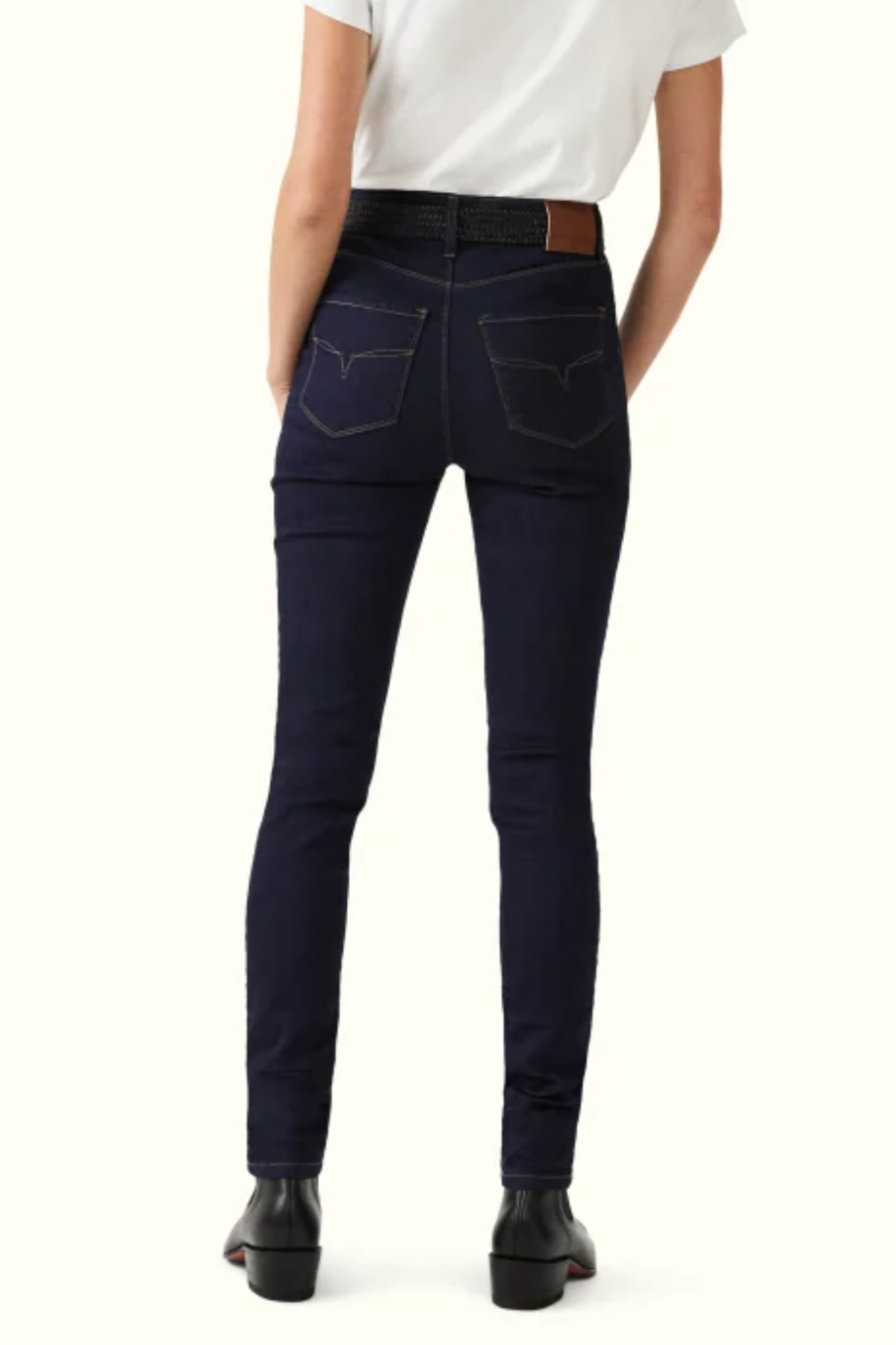 A person standing with their back facing the camera, wearing a white t-shirt, RM Williams Albury Jean with dark high-rise skinny fit, visible pocket stitching and Longhorn embroidery, a black belt with brown leather detail, and black boots. The background is light beige.