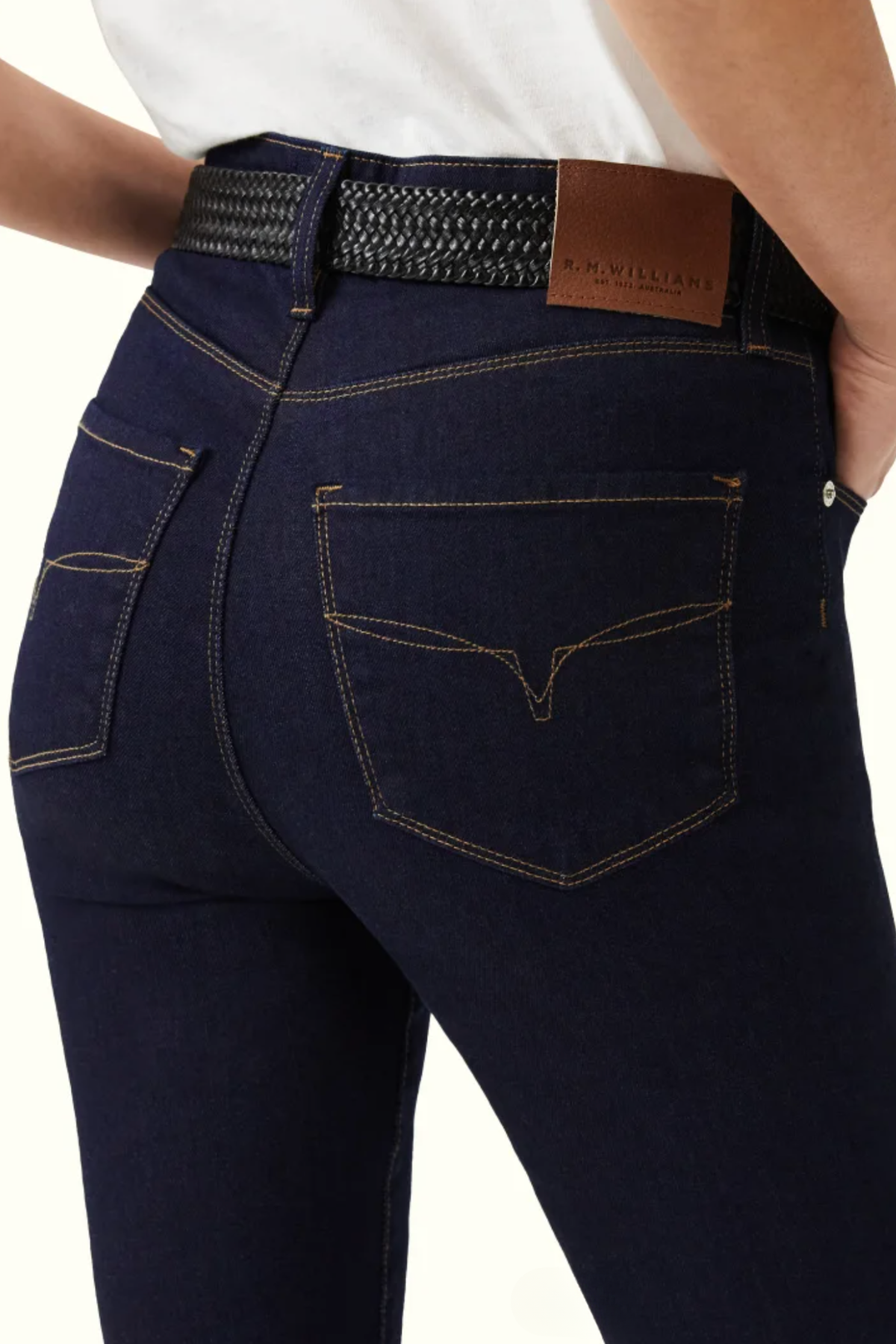 Close-up view of a person wearing the RM Williams Albury Jean, which are high-rise skinny fit jeans made from stretch cotton. The jeans are paired with a white shirt and a black woven belt. They feature brown stitching and have a brown leather patch on the belt with the brand "R.M. Williams" embossed on it.