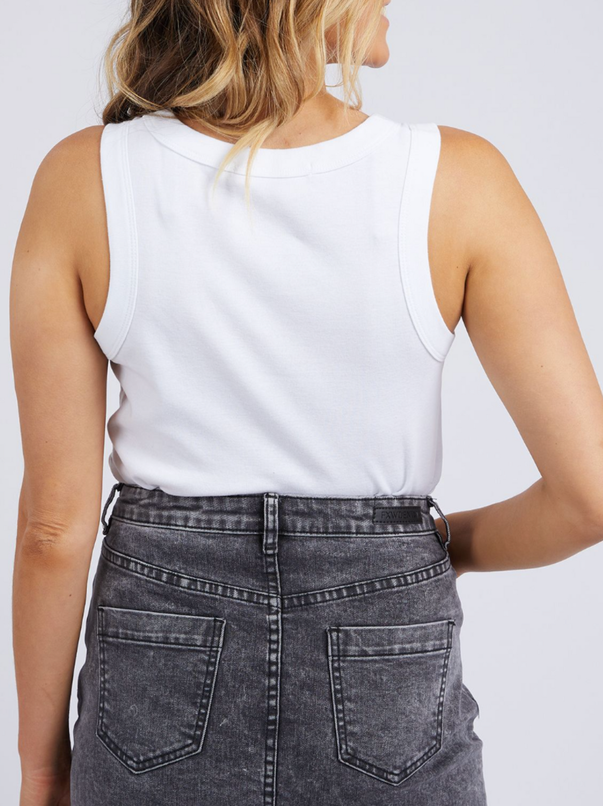 A woman is seen from behind in the Foxwood Esme Tank, a white rib knit sleeveless top, paired with a high-waisted faded black denim skirt with back pockets. Her light brown wavy hair cascades partially down her back.