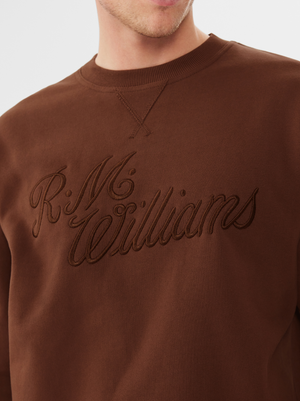 A person wearing the RM Williams R.M.W Script Crew Neck—a brown cotton blend jumper with matching thread "R.M.Williams" embroidery on the front. Only the lower half of their face, neck, and upper torso are visible.