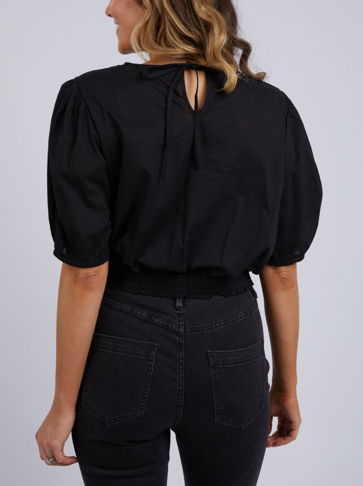 The image showcases a person with wavy hair wearing the Foxwood Charli Top, a black blouse with short puff sleeves and a tie at the back. They're paired with high-waisted dark cropped jeans, turned away and visible from shoulders to hips.