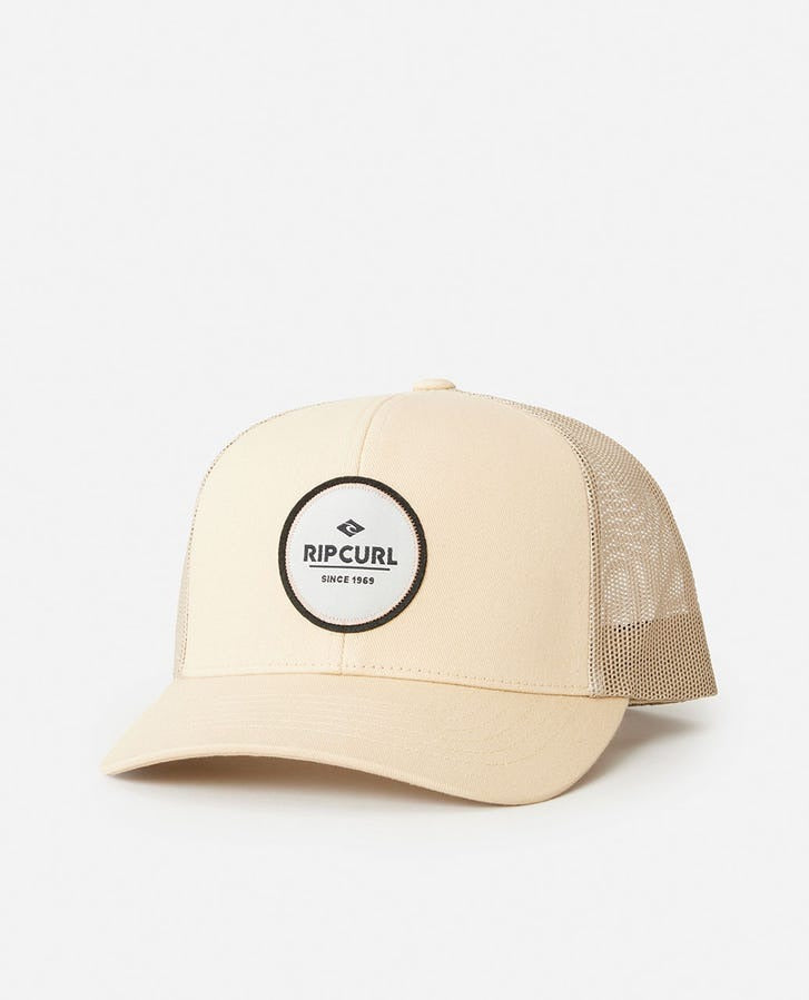 The Rip Curl Routine Curve Trucker is a beige hat with a mesh back and a round black-and-white Rip Curl logo patch on the front, shown against a white background.