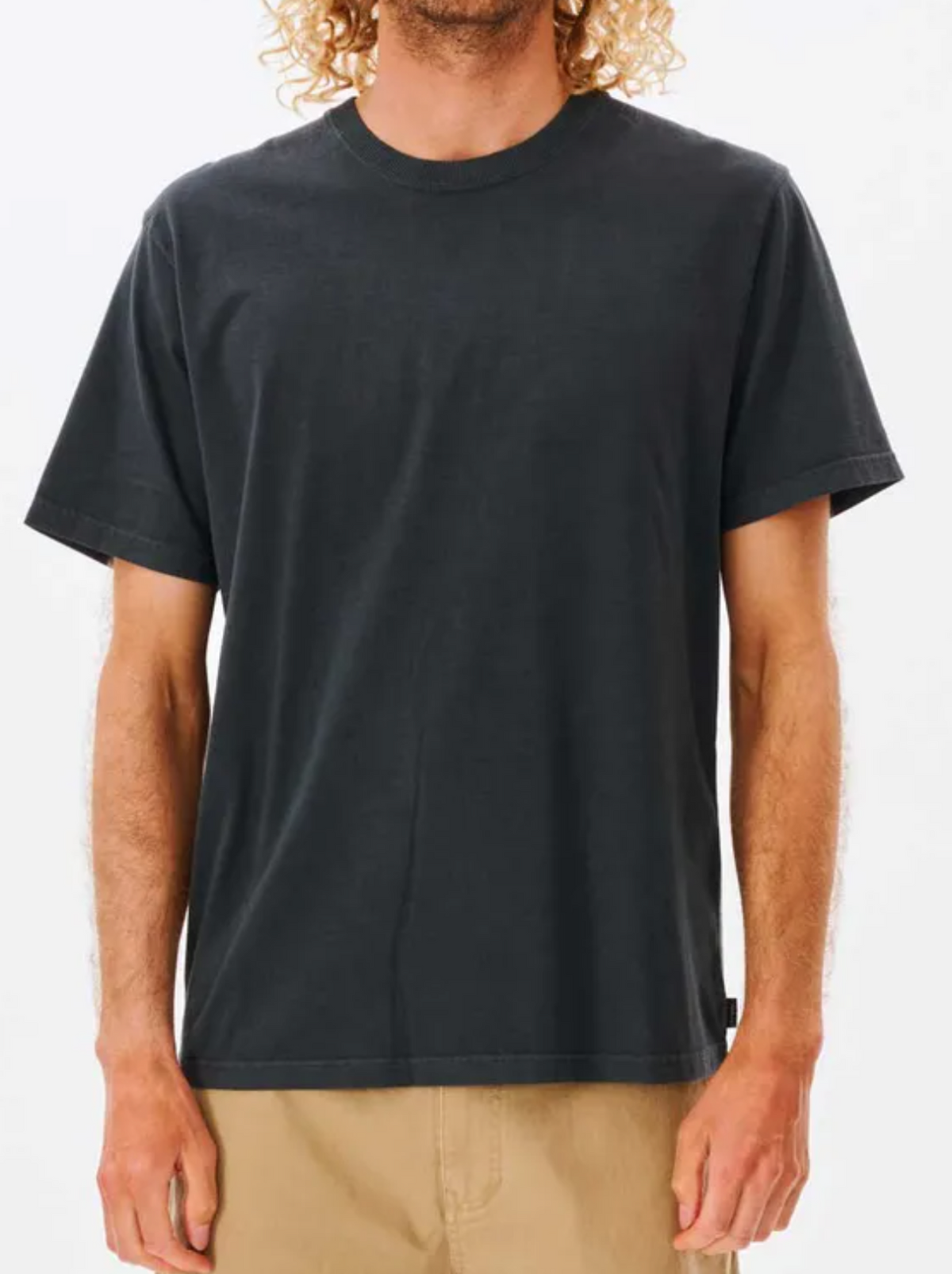 A person with curly hair models the Rip Curl Plain Wash Tee in black with tan pants, standing before a white background. The relaxed fit of the tee is visible, showing their torso and part of their face.