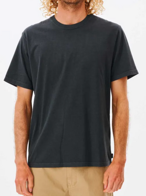 A person with curly hair models the Rip Curl Plain Wash Tee in black with tan pants, standing before a white background. The relaxed fit of the tee is visible, showing their torso and part of their face.