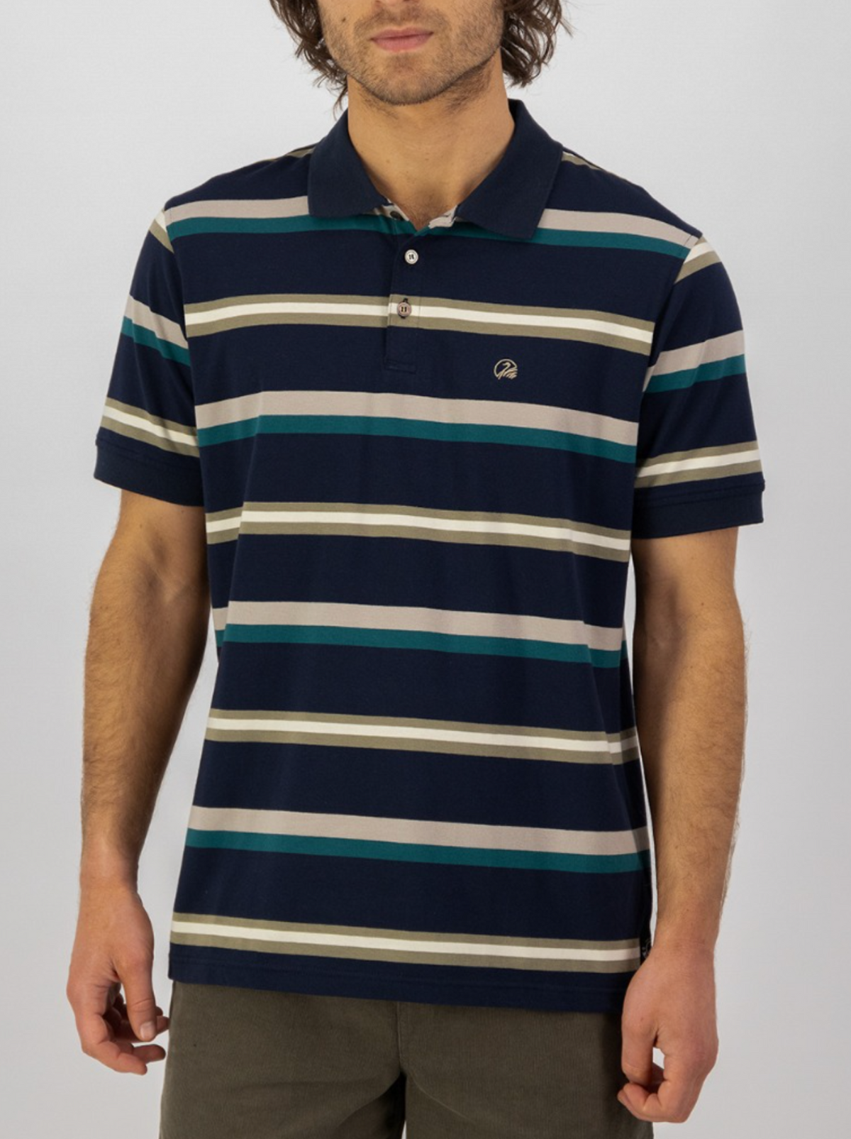 A man wears the Swanndri Winfred Stripe Polo, a cotton-elastane blend top, with dark green pants. He stands against a plain light background, head slightly tilted and arms at his sides.