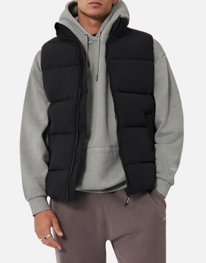 Someone is wearing a gray hoodie layered under an Industrie The New Hanam Puffer Vest with quilted padding, paired with dark gray pants. Their hand is partially visible at their side against a plain white background.
