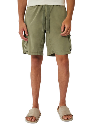 A person is seen wearing Industrie The Del Sur Combat Short in olive green, made of 100% cotton with multiple side pockets, and beige slippers against a plain background. Only the lower half of the body is visible.