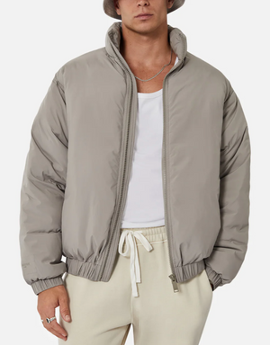 A person wears the Industrie The Ottawa Puffer Jacket in light gray over a white tank top and cream drawstring pants, styled with a silver chain necklace and gray bucket hat. The background is plain white.