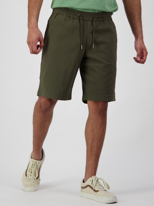 A person wearing Swanndri Clevedon Stretch Canvas Shorts in olive green, a light green shirt, and white sneakers with brown accents stands against a plain white background, with their hands relaxed by their sides.