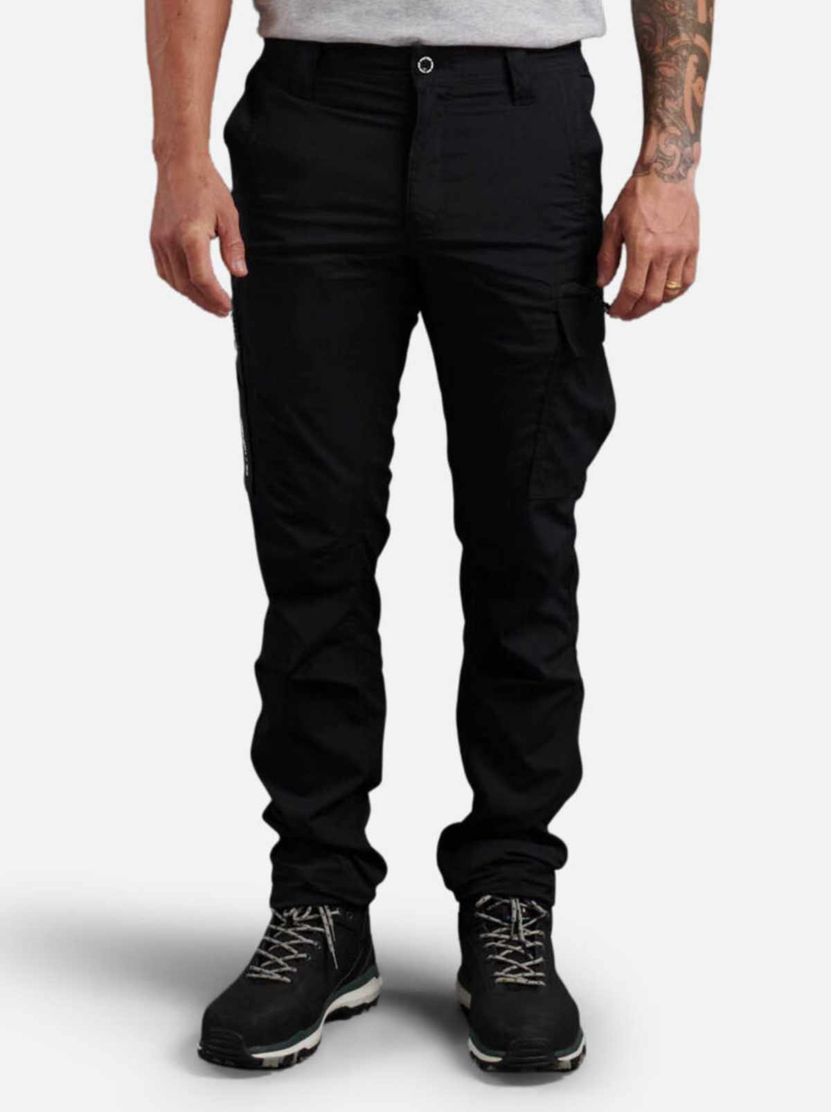 A person with a tattooed arm stands against a plain white background, wearing black King Gee Trademark Cargo Pants, black hiking boots, and a light grey T-shirt.