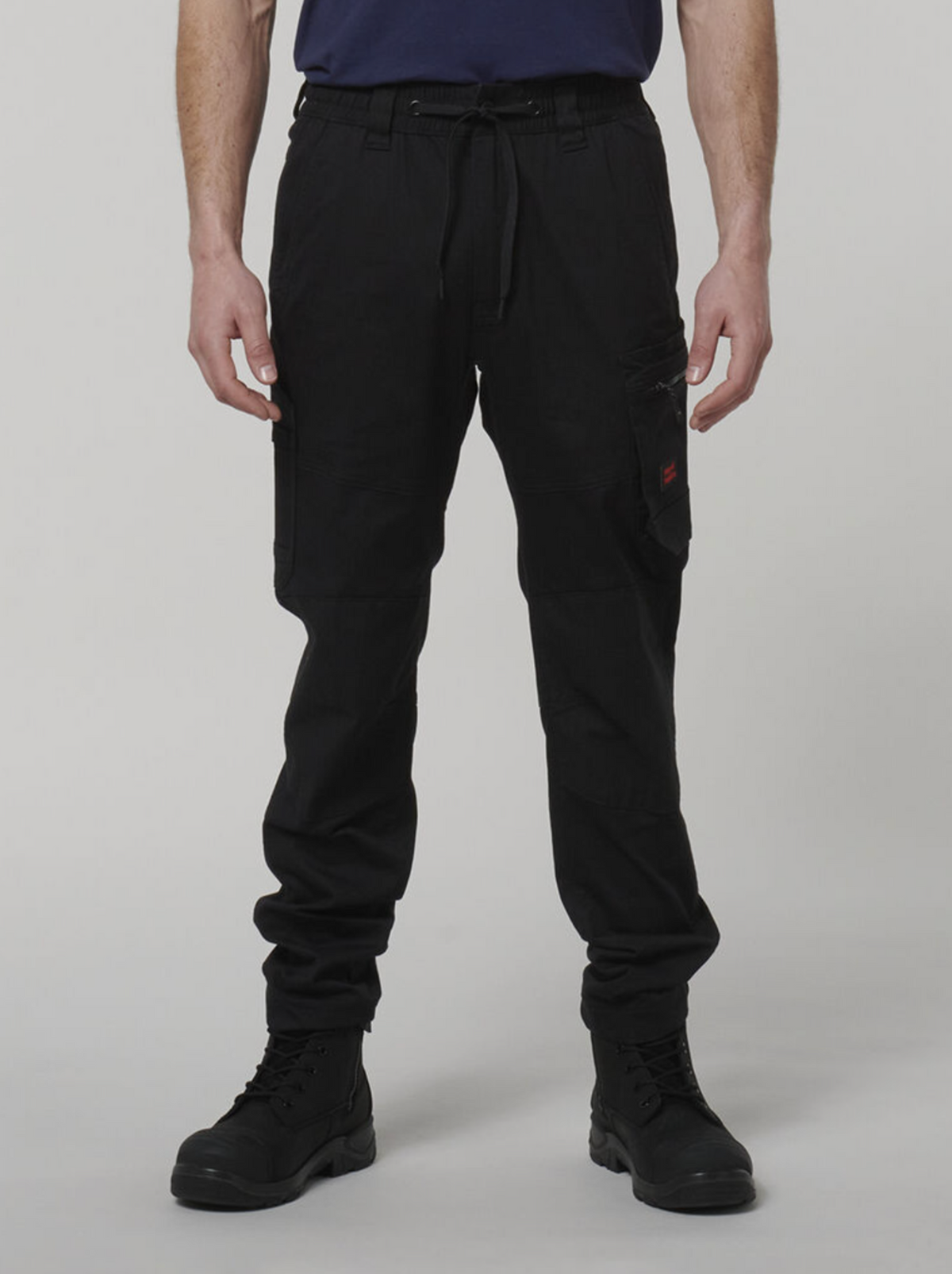 A person wearing black Hard Yakka Tough Maxx Pant cargo pants, black boots, and a navy blue shirt stands with arms relaxed by their sides against a plain light background.