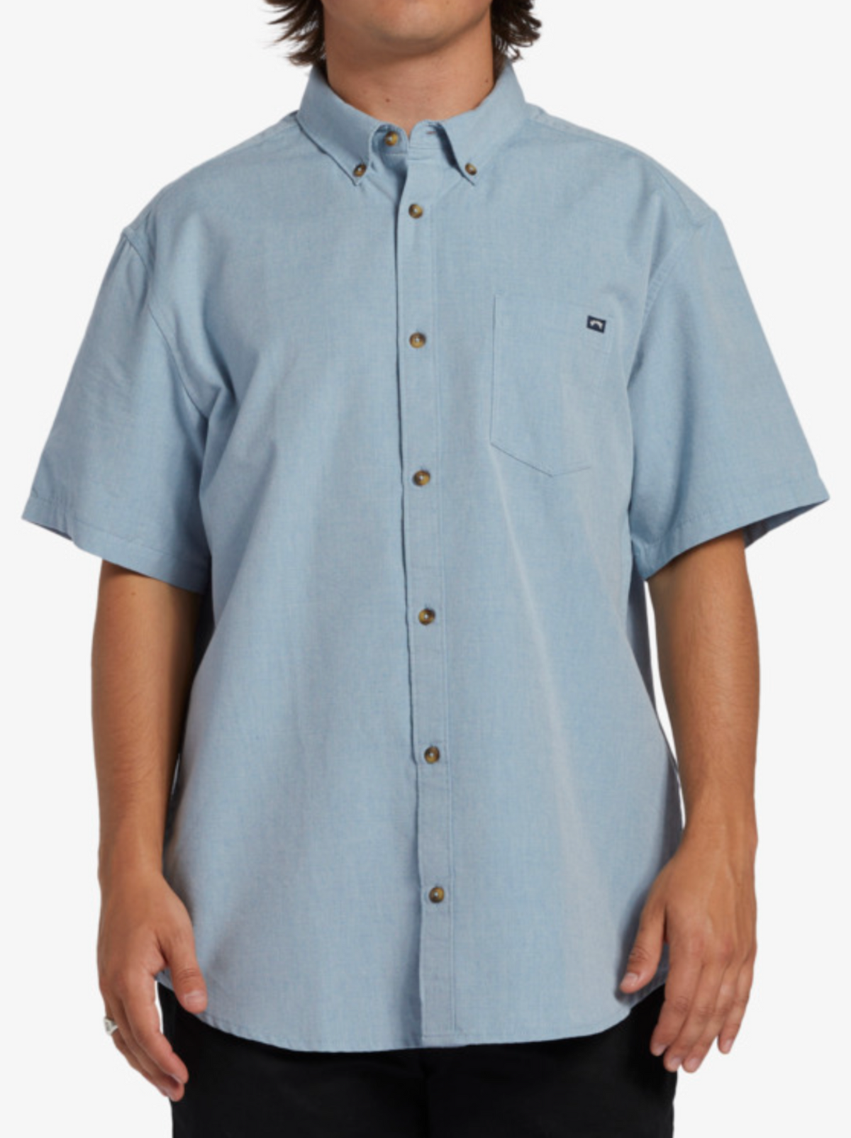 A person wears the Billabong All Day Short Sleeve Shirt in light blue, featuring soft woven fabric, a chest pocket, and a small logo tag, standing against a plain white background—a nod to classic roots.