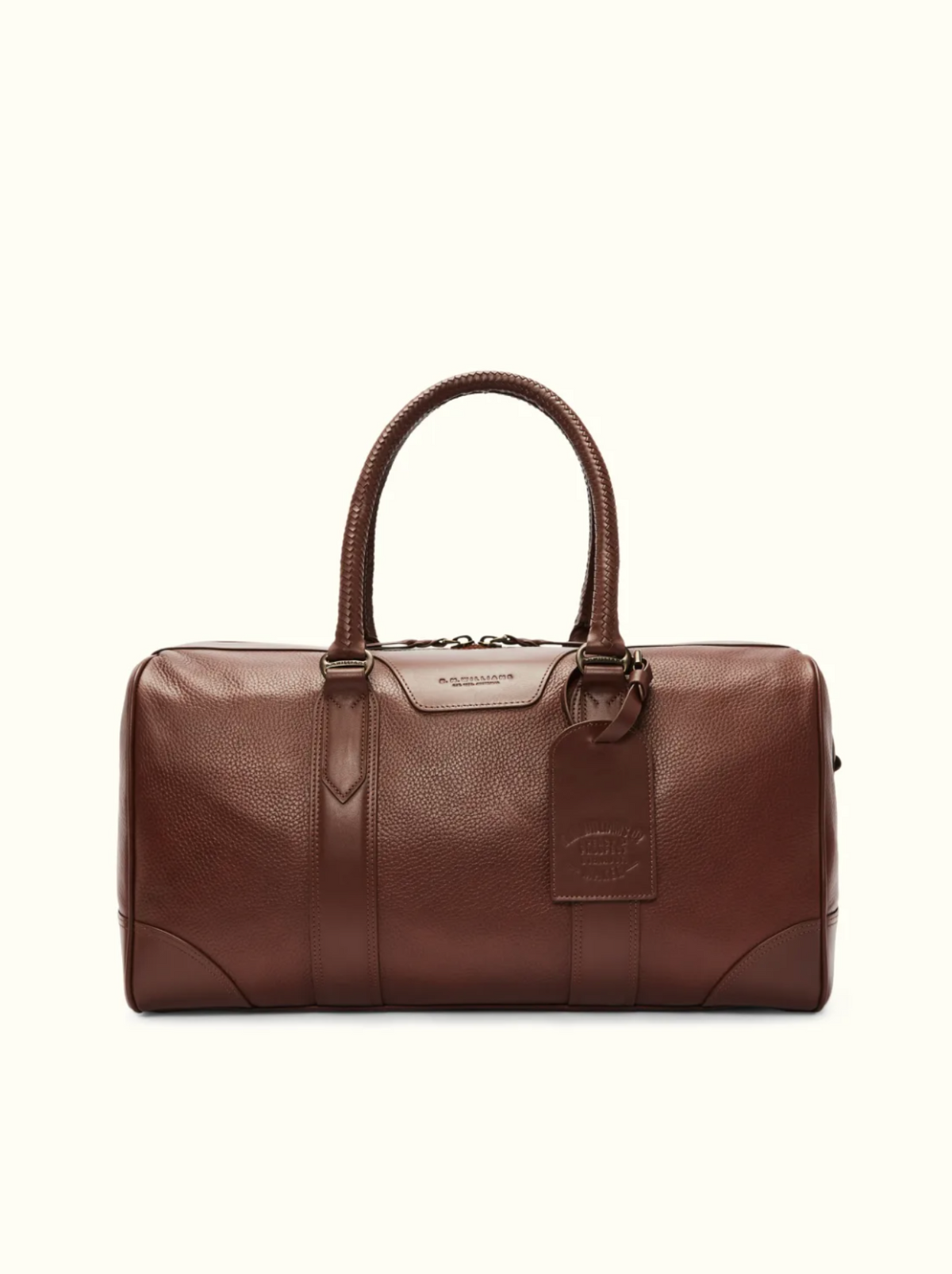The RM Williams Overnight Bag is a brown full grain leather duffel with a pebbled texture, rounded handles, luggage tag, metal hardware, and top zipper closure. It's set against an off-white background to complement your travel items.