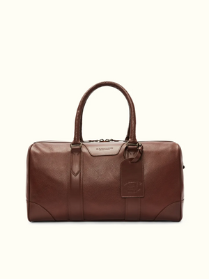 The RM Williams Overnight Bag is a brown full grain leather duffel with a pebbled texture, rounded handles, luggage tag, metal hardware, and top zipper closure. It's set against an off-white background to complement your travel items.