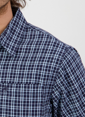 A close-up shows someone wearing a Swanndri Russell Shirt in blue and white checks, highlighting the pointed collar and chest pocket. Made from 100% cotton with a relaxed fit, it is visible from neck to upper chest against a plain light background.