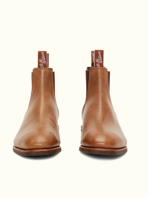A pair of RM Williams Comfort Craftsman Boots viewed from the front. The brown leather boots feature a smooth surface with elastic side panels, chisel square toes, and pull tabs on the back displaying branding text. The darker brown soles contrast against a plain, light-colored background.