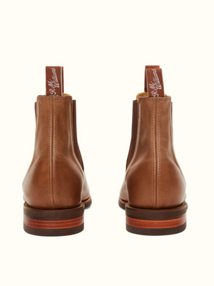Back view of a pair of RM Williams Comfort Craftsman boots, showcasing the brown leather Chelsea design with pull tabs displaying the brand name. These sleek boots feature stitched details, a chisel square toe, and a wooden sole with a rubber heel, all against a plain background.