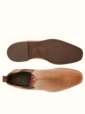 The RM Williams Comfort Craftsman Boot in brown leather is displayed from two perspectives: a top-down view of one boot and a bottom view showing the sole. The boots feature pull tabs at the back and sleek, chisel square toes. The sole has a textured pattern for grip and bears a logo imprint.