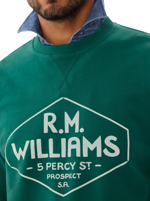 A man wears the RM Williams Gladstone Crew Neck—a relaxed-fit, cotton fleece green sweatshirt with a white “5 Percy St Prospect S.A.” graphic—over a blue collared shirt, with only the lower half of his face visible.