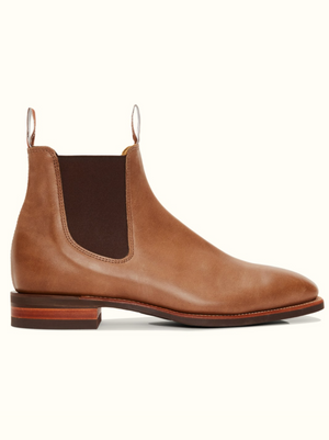 A single RM Williams Comfort Craftsman Boot in tan leather with a chisel square toe, featuring a brown elastic side panel, a pull tab at the back, and a wooden sole with a low heel, viewed from the side on a plain white background.