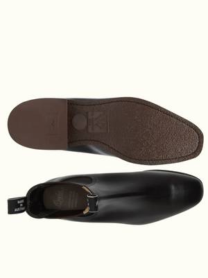 A pair of RM Williams Comfort Craftsman Boots in black is shown. One boot is positioned to display the top view and side profile, emphasizing the smooth pull-up leather surface and elastic side panel. The other boot showcases the durable rubber sole, featuring a textured brown surface with branding, ideal for modern comfort.