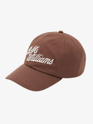 The RM Williams Script Cap is a brown cotton twill baseball cap with "J.M. Williams" embroidered in white cursive on the front, featuring an adjustable strap and shown on a plain white background.