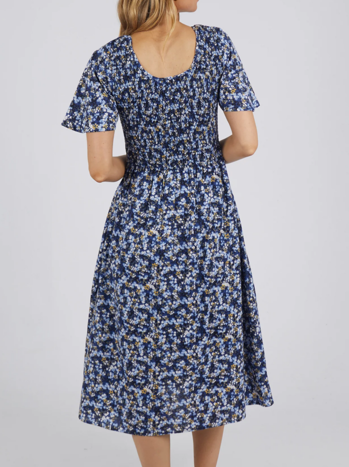 A woman with blonde hair models the Foxwood Lulu Dress in sapphire ditsy print, made from cotton poplin. She faces away, showing the short-sleeved, calf-length design with a shirred bodice against a plain light background.