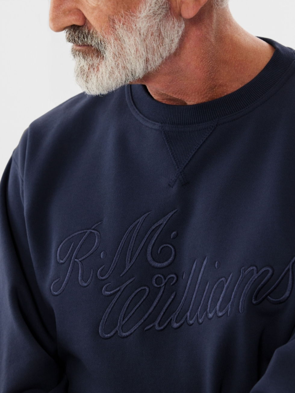An older man with a white beard wears the navy RM Williams R.M.W Script Crew Neck, a soft cotton blend jumper featuring an embroidered "R.M. Williams" logo on the front, partially revealing his profile.
