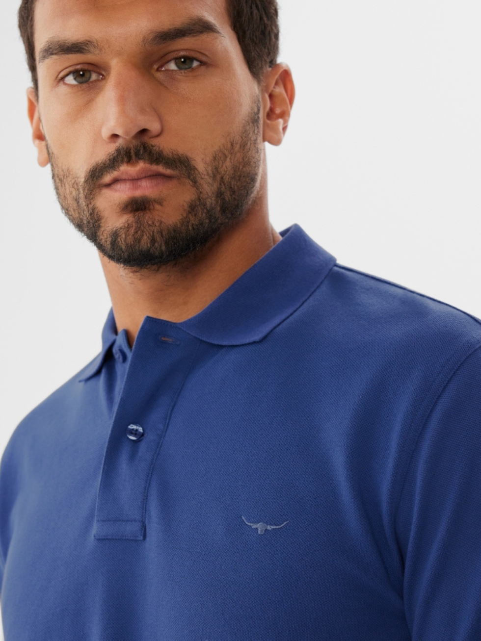 A man with a short beard and mustache wears the RM Williams Rod Polo, a blue cotton pique shirt with a small longhorn embroidery on the chest, looking directly at the camera against a plain light background.