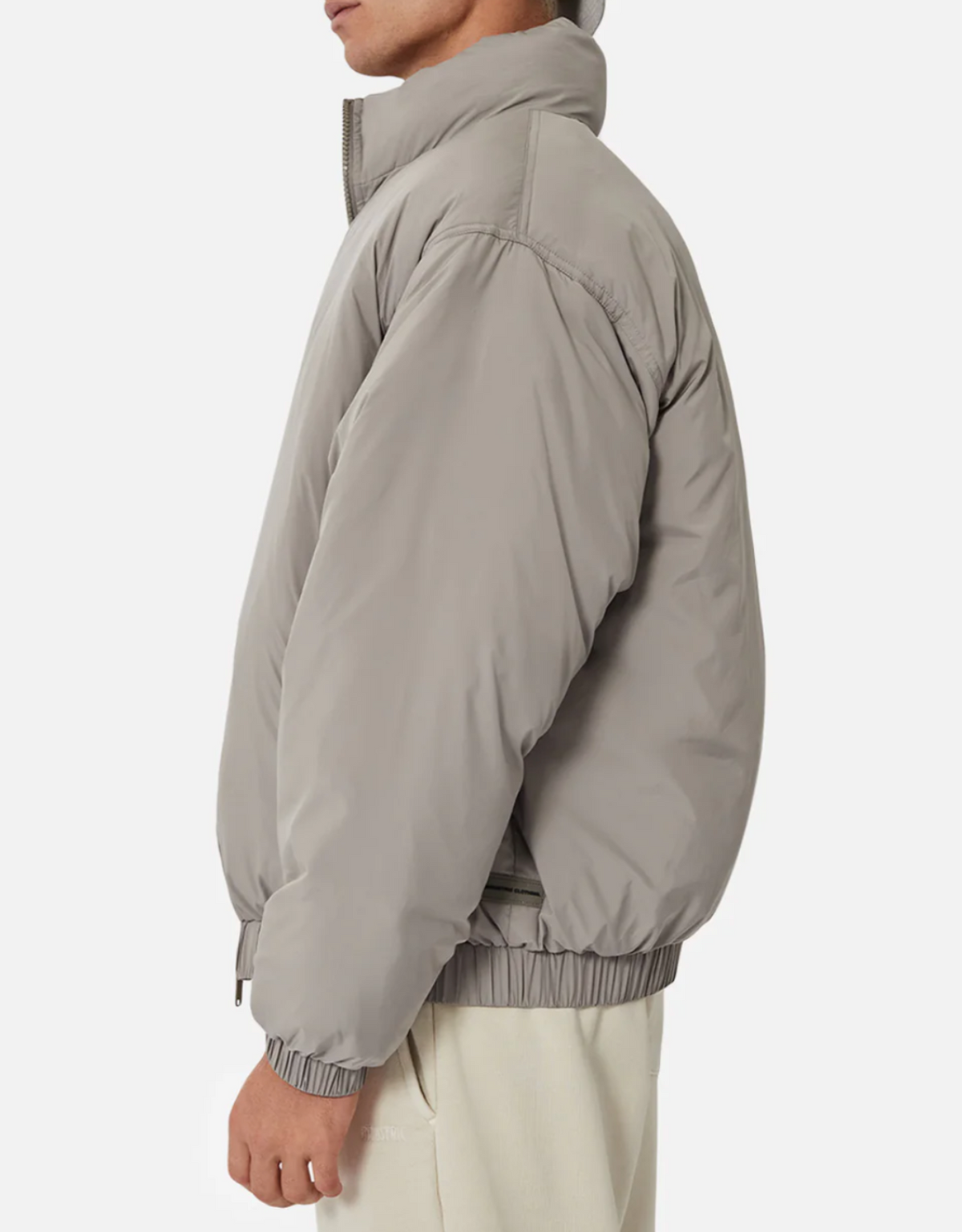 A person stands sideways against a plain white background, wearing the Industrie The Ottawa Puffer Jacket in light gray with a high collar, elastic hem and cuffs, styled with light-colored pants for a modern, minimalist look.