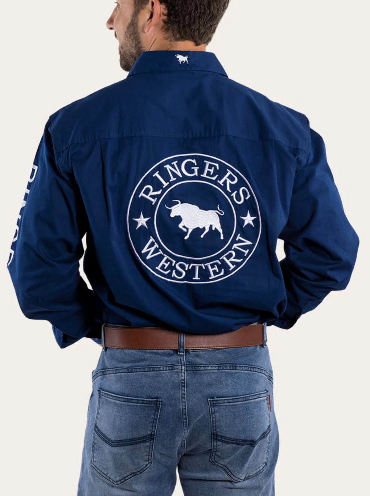 A man seen from behind wears a blue Ringers Western Hawkeye Embroidered Shirt with UPF50+ protection, featuring a large white "Ringers Western" logo and bull graphic on the back, paired with jeans and a brown belt.