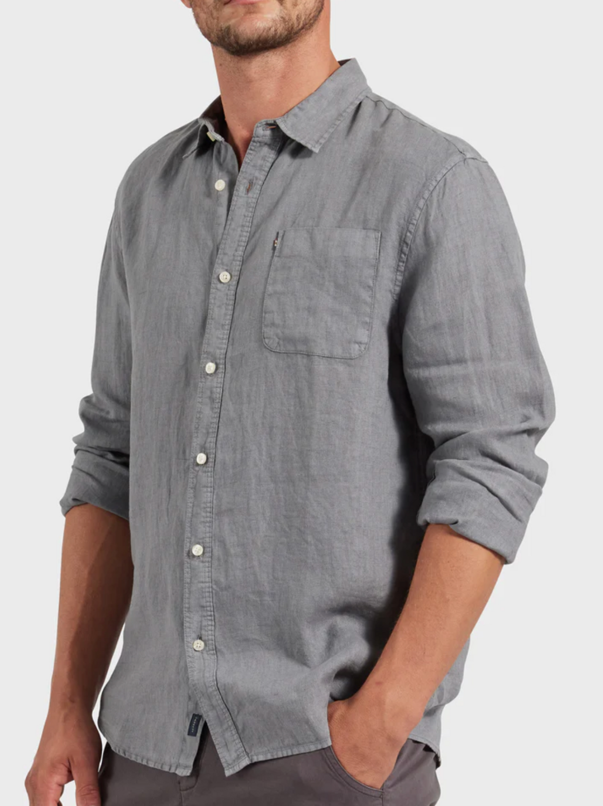 A stylish man wearing the Academy Brand Hampton Linen Shirt in light gray with rolled-up sleeves stands with one hand in his pocket against a plain light background.