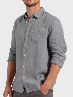 A stylish man wearing the Academy Brand Hampton Linen Shirt in light gray with rolled-up sleeves stands with one hand in his pocket against a plain light background.