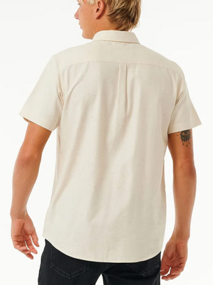 A person with short blond hair is wearing a Rip Curl Ourtime Short Sleeve Shirt in light beige and black pants, facing away from the camera. A tattoo is visible on their left arm.