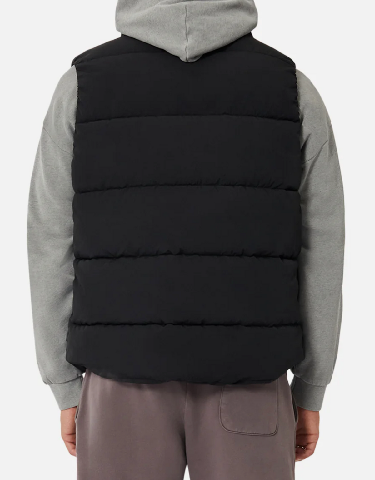 A person wearing an Industrie The New Hanam black puffer vest with quilted padding over a gray hoodie is seen from behind, paired with brown pants against a plain white background.